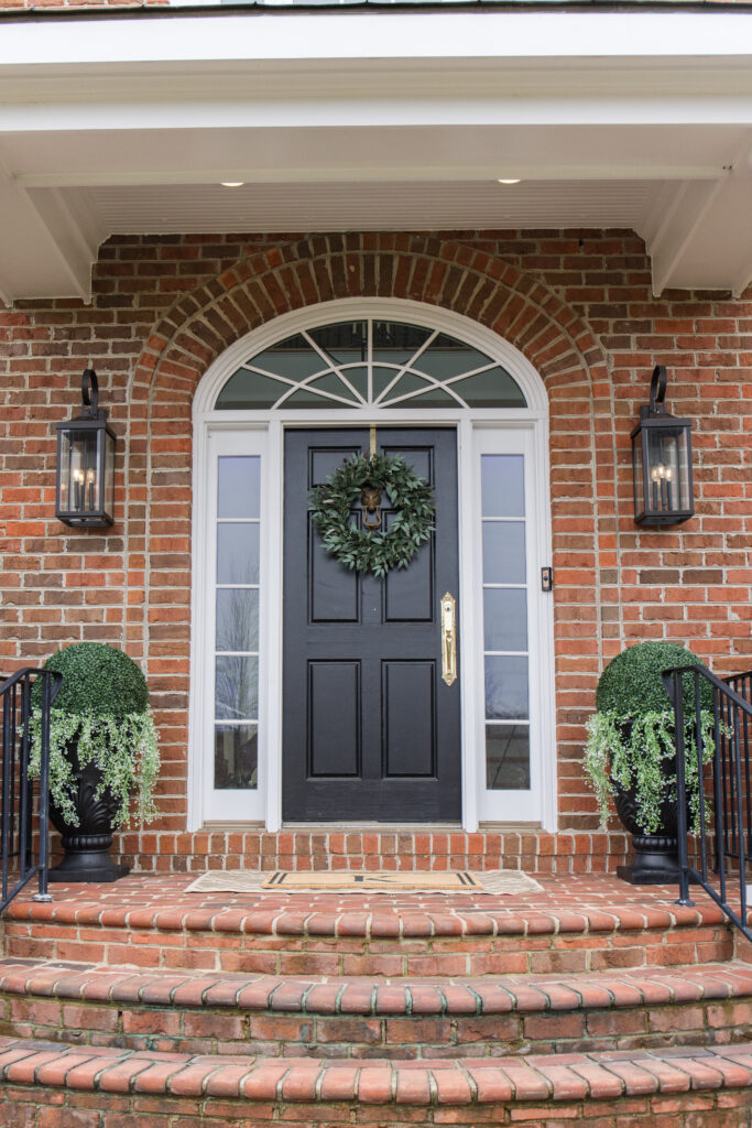 arched manor spring front porch styling, image of front porch in real life with black door, gold handles, wreath, sconces, shrubs, planters, doormat, and neutral rug
