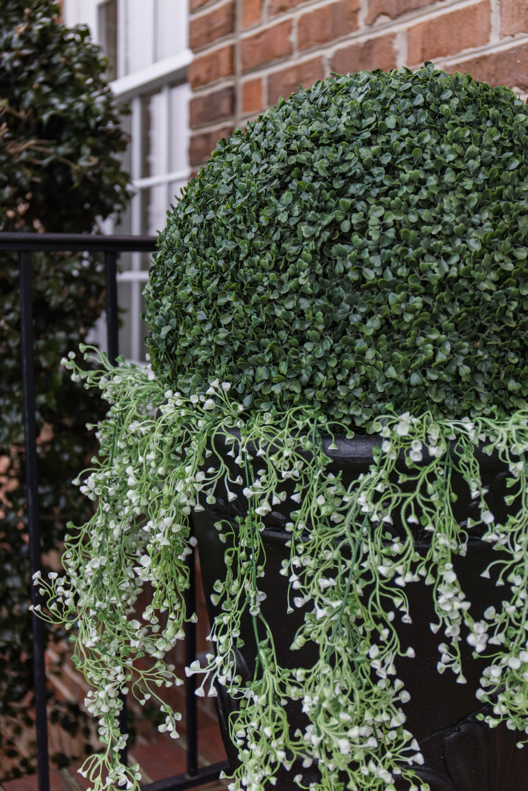 Arched manor spring front porch decor, close up image of large faux shrubs ball styled with hanging plants in black decorative planter