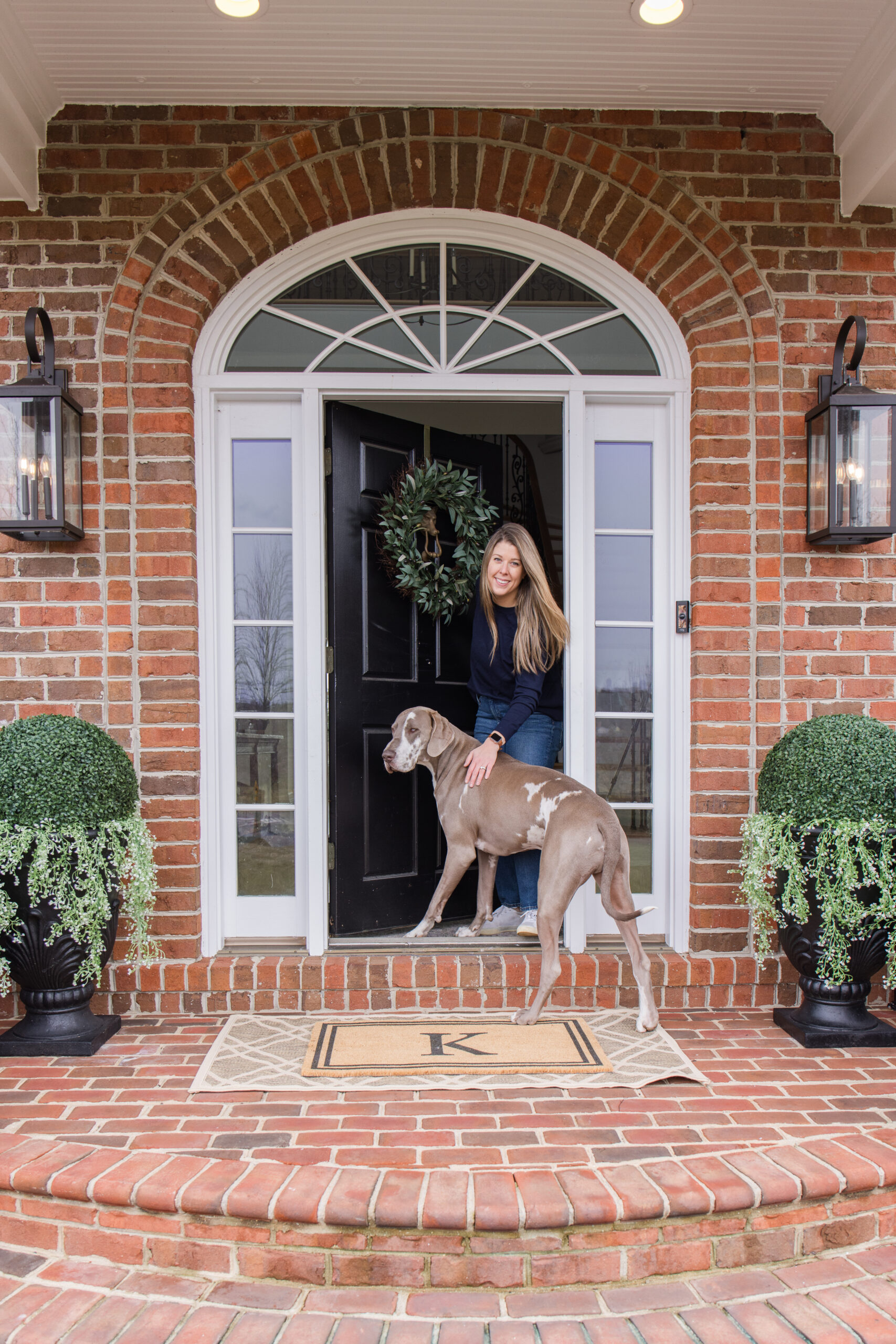Spring front porch styling Arched Manor spring front porch styling featuring Megan and dog Slinky | inital doormat | outdoor rug | large shrubs styling in black planter with hanging plants | black wall sconce | olive wreath on front door