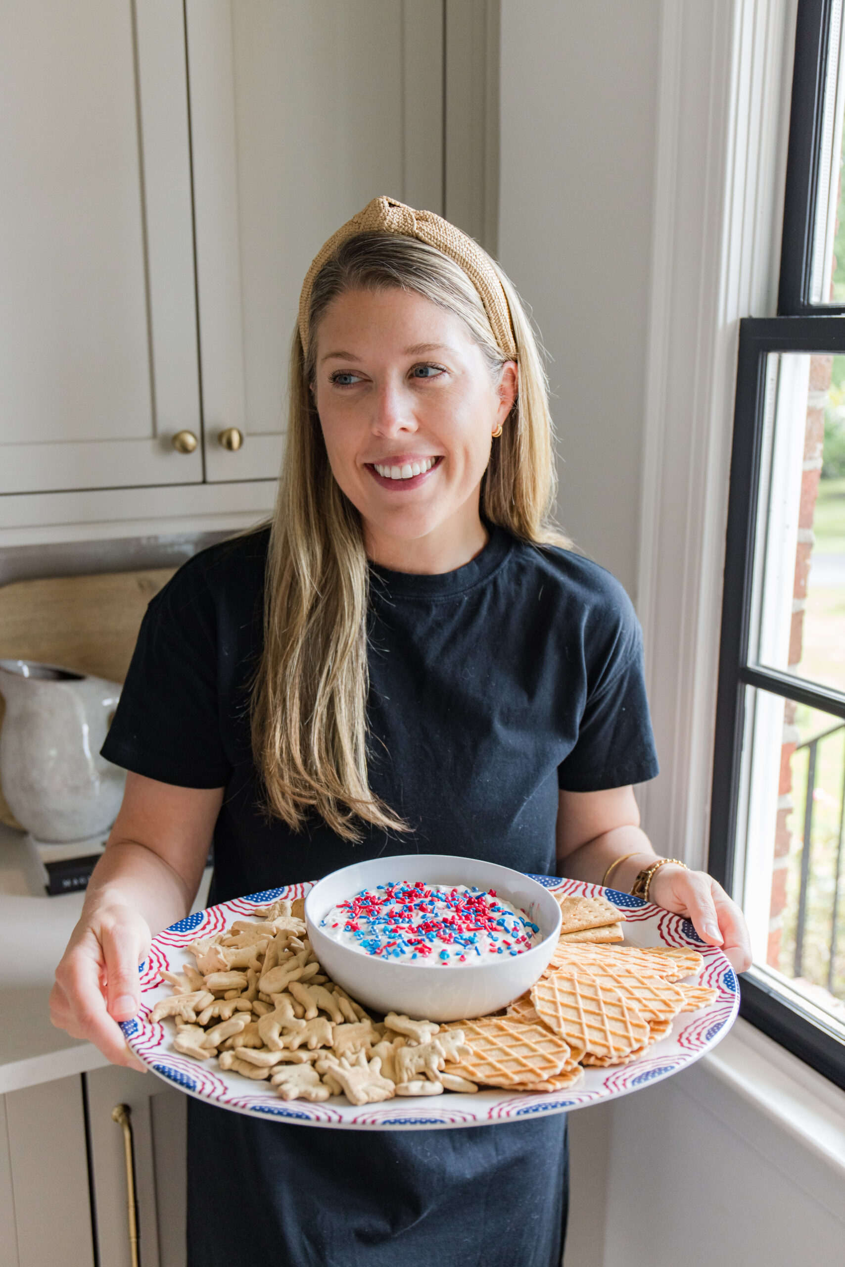 megan wearing a navy blue dress holding a serving platter with snacks and a white bowl filled with dip