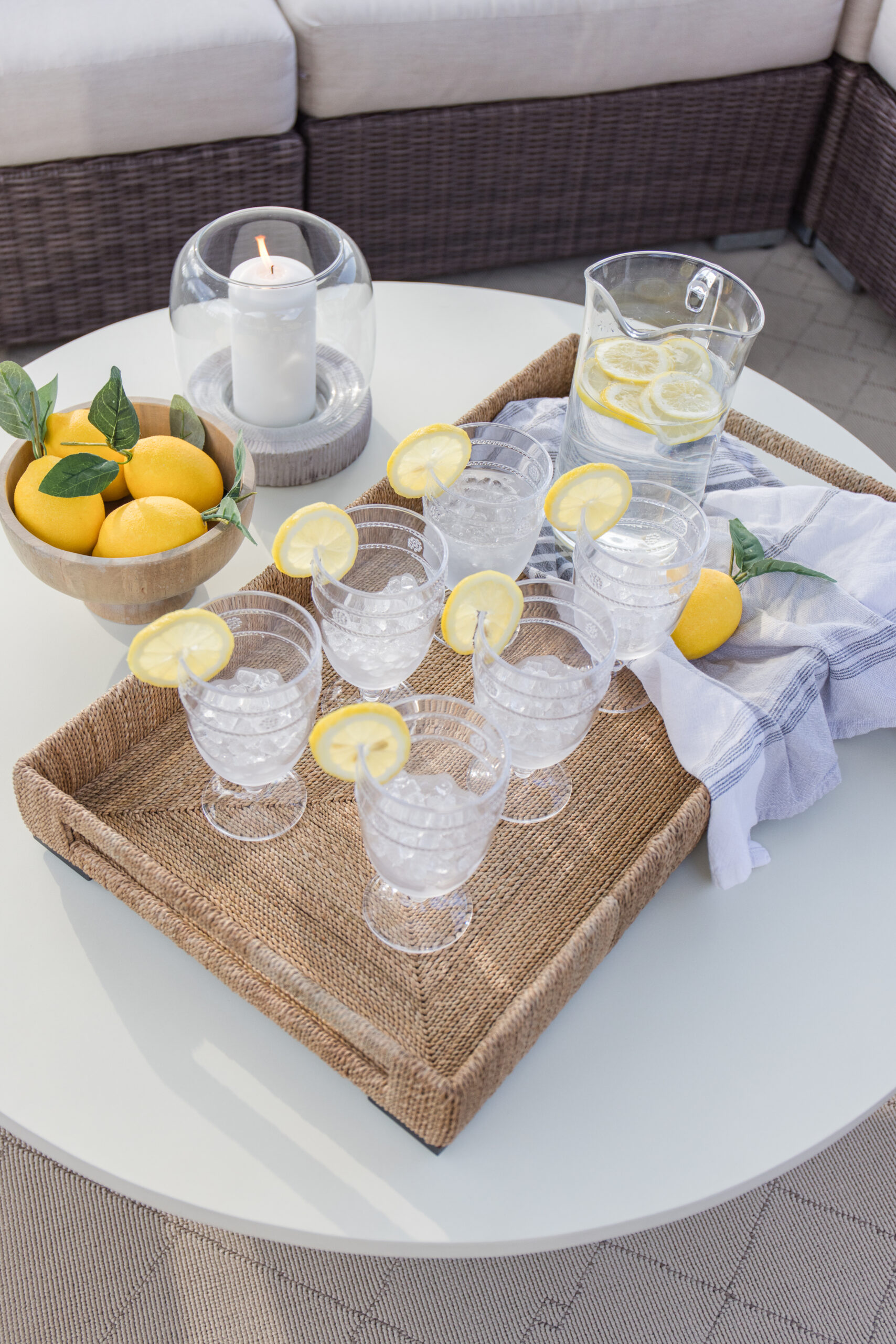 woven serving tray with goblet glasses filled with ice and garnished with a lemon slice, small decorative bowl filled with faux lemons, a hurricane candleholder, a pitcher of lemon water